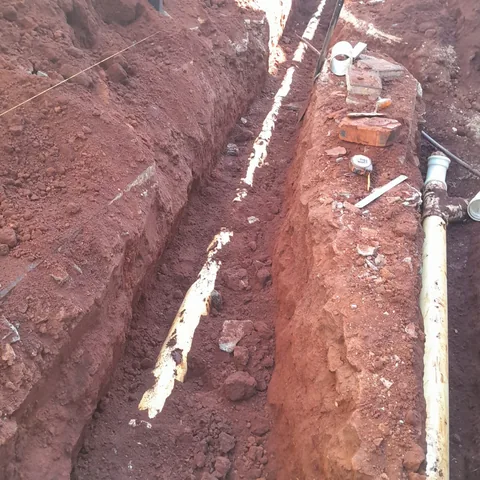 White PVC conduit laid in a deep trench in red soil with measuring tools on the bank